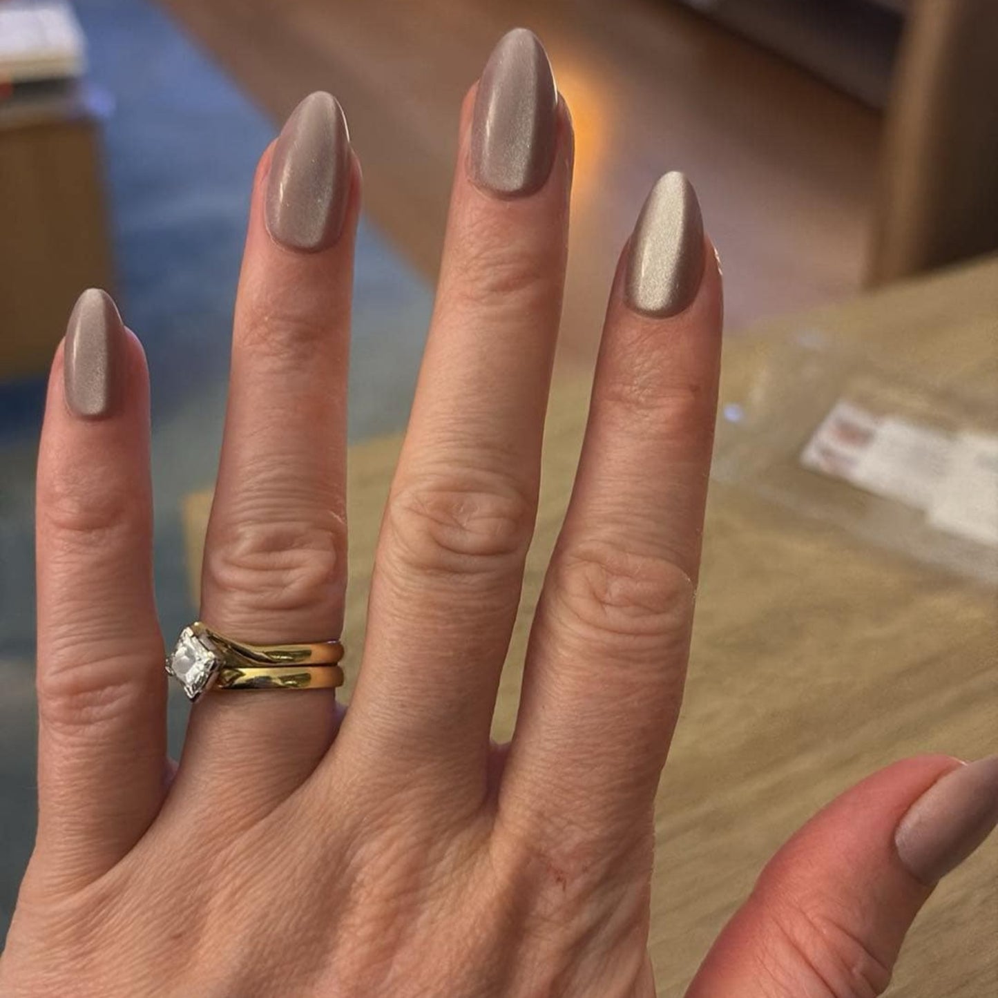Left hand wearing almond shaped nails in a shiny chocolatey colour, with a lounge room in the background.