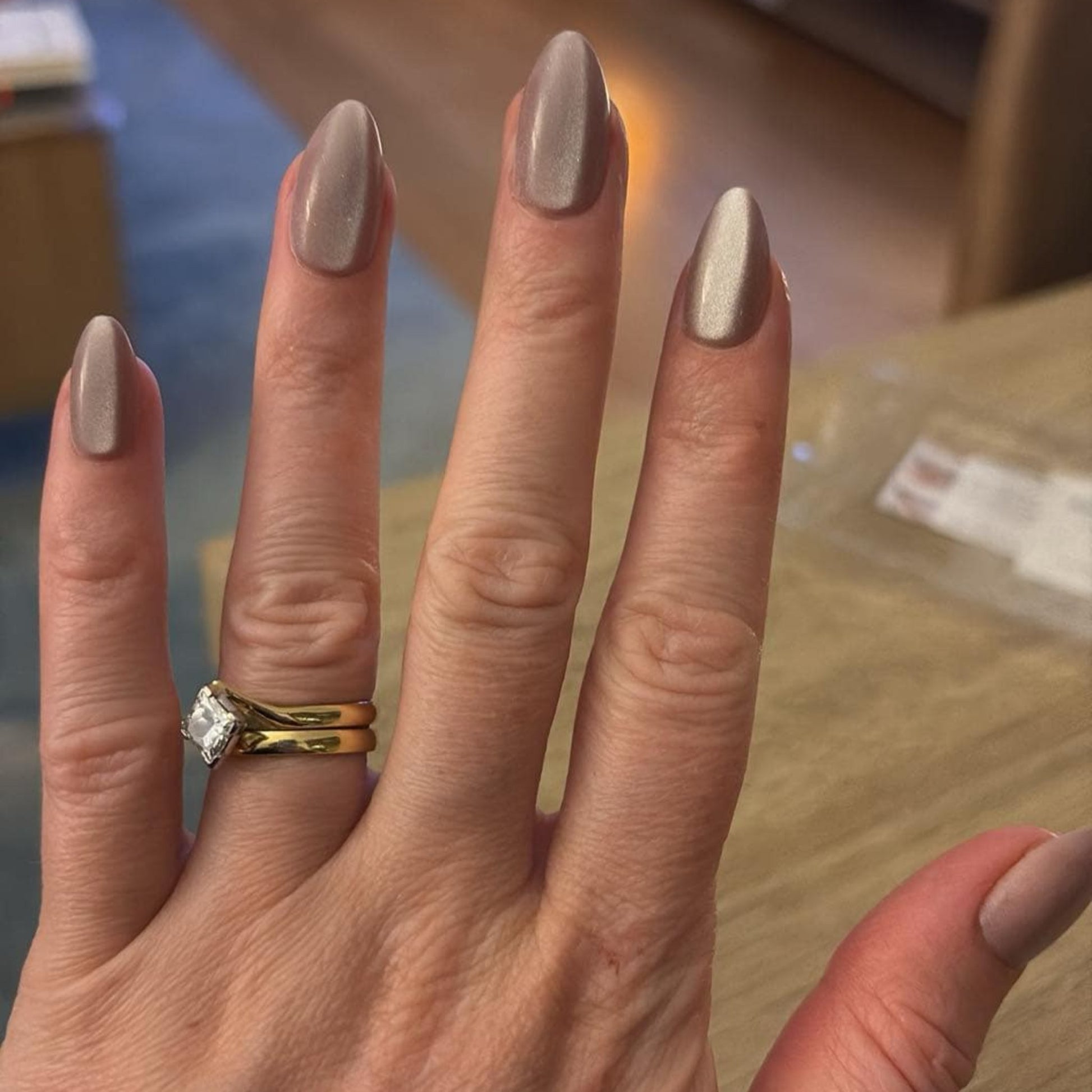 Left hand wearing almond shaped nails in a shiny chocolatey colour, with a lounge room in the background.