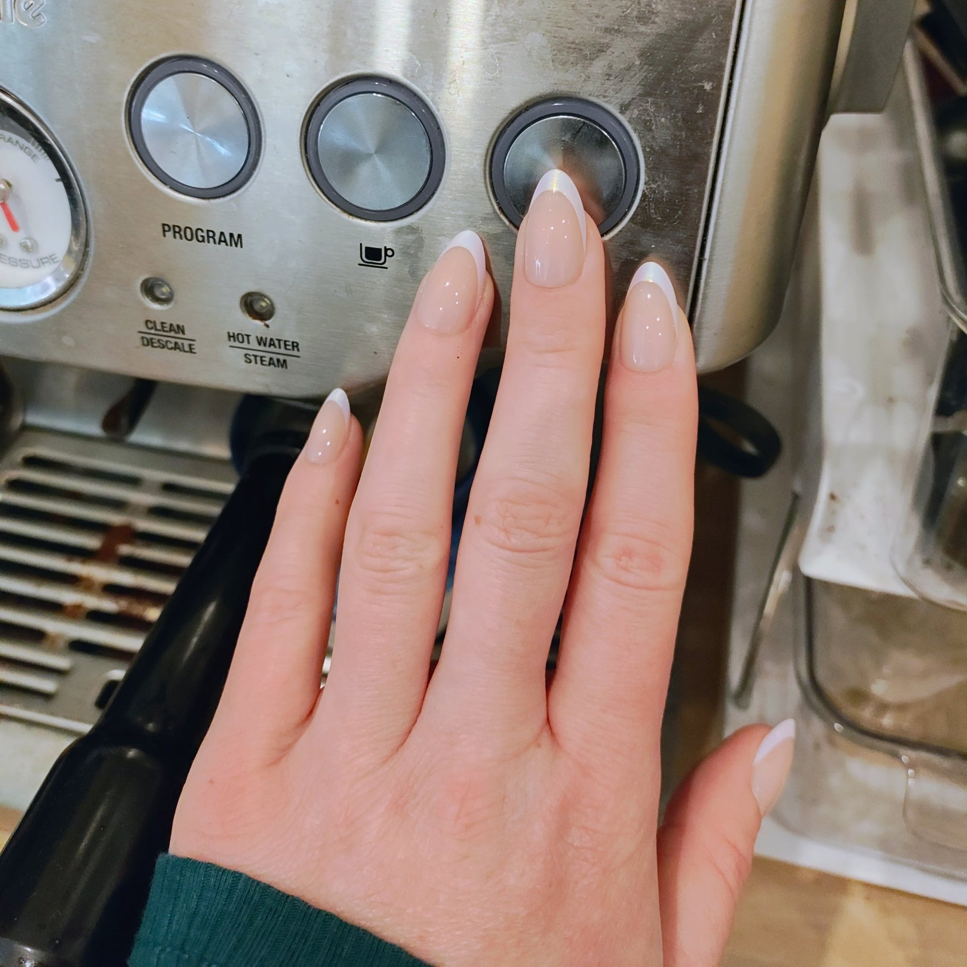 Left hand with almond frosted French tip nails pressing a button on a coffee machine.