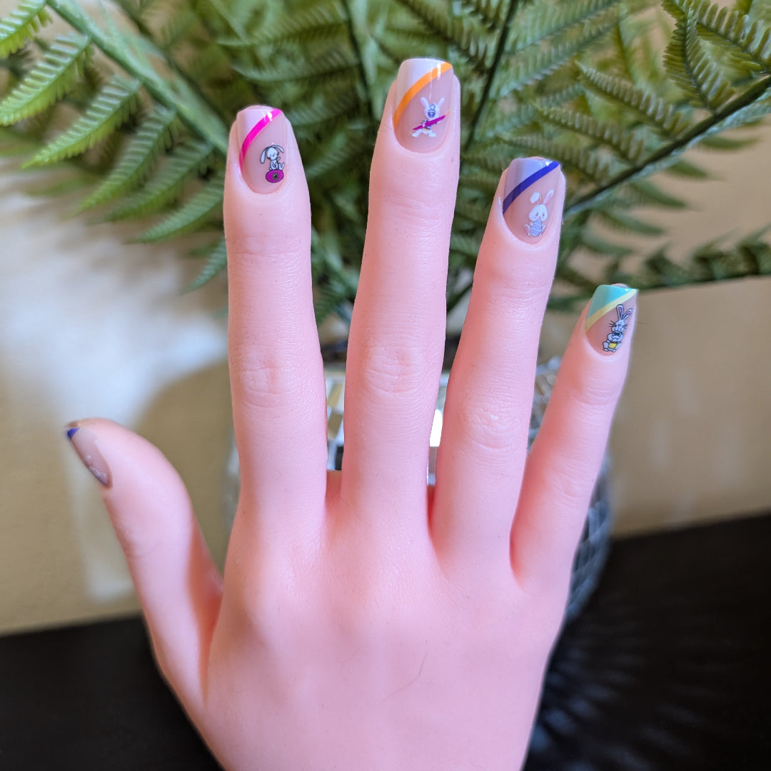 A right hand with square sherbert nails with Easter Bunny designs behind a fern.