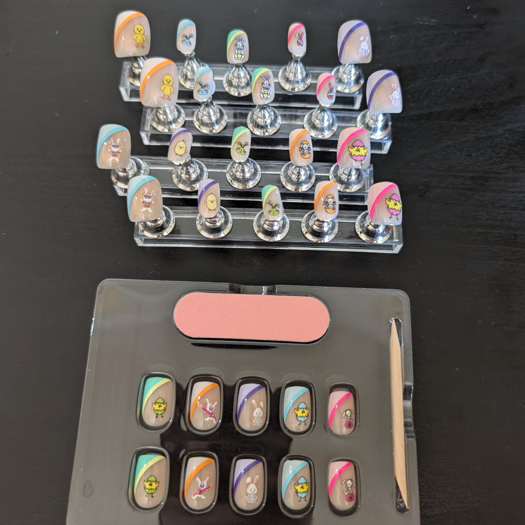 20 square nails of varying sizes that have easter bunny and chick details with swatches of colour and a box of 10 nails with a cuticle stick and nail file underneath.