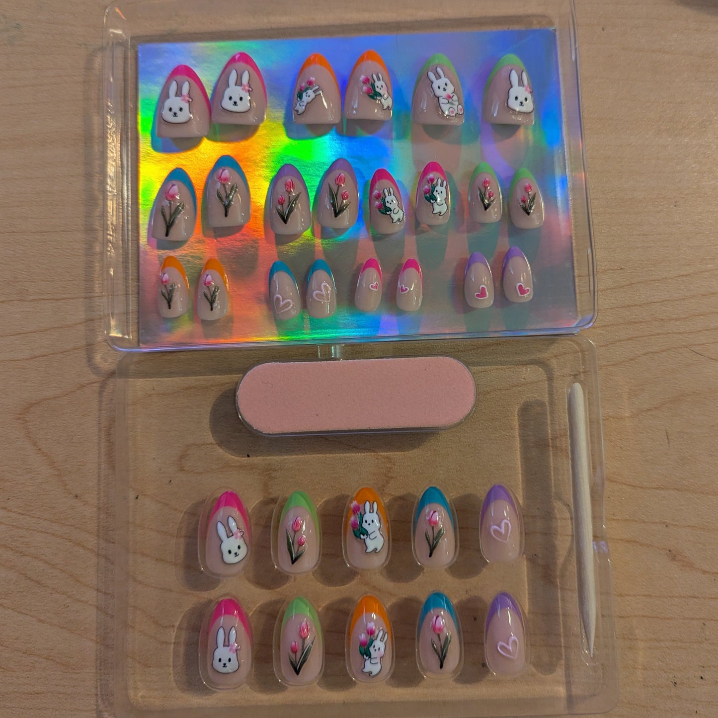 Image of a clear box with 10 almond press on gel nails with various tip colours and Easter designs, a cuticle stick and a nail file. On top there is another clear box with almond gel press-ons with 22 nails of varying tip colours and Easter designs.