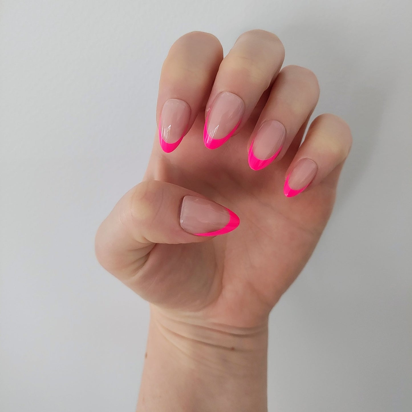 Left hand showing almond nails with a hot pink French tip on a white wall background