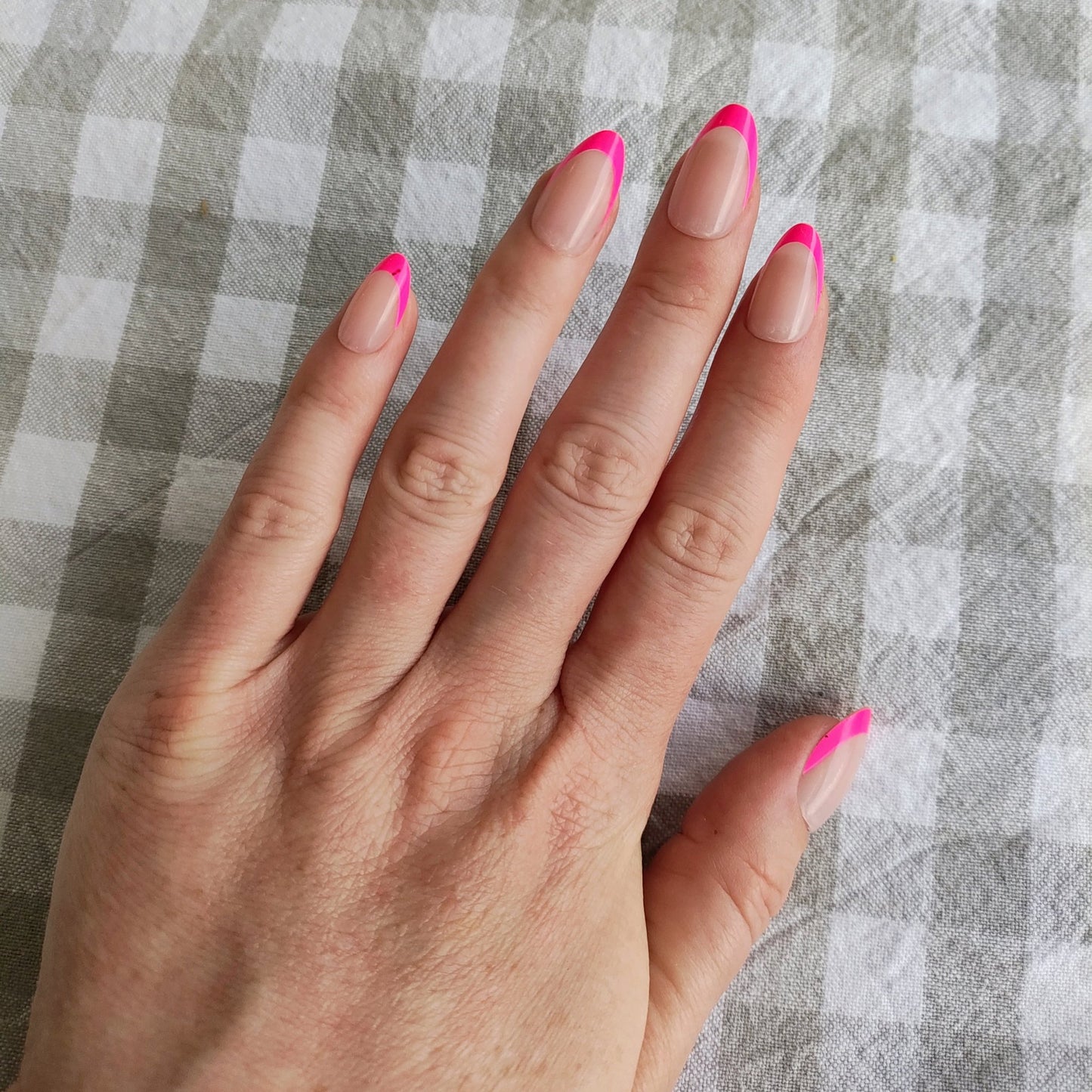 Left hand showing almond nails with a hot pink French tip on a grey Gingham tablecloth