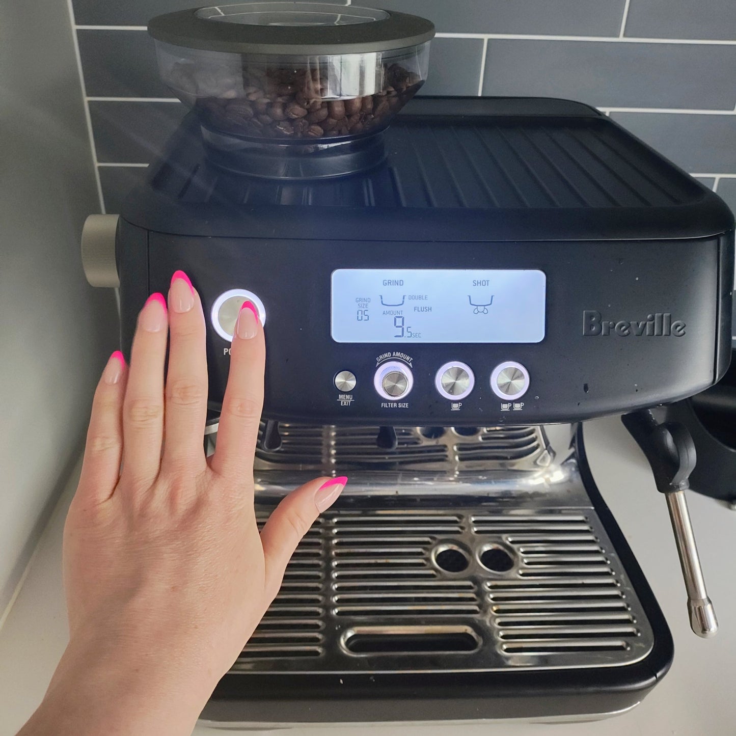 Left hand showing almond nails with a hot pink French tip turning on a black home espresso machine.