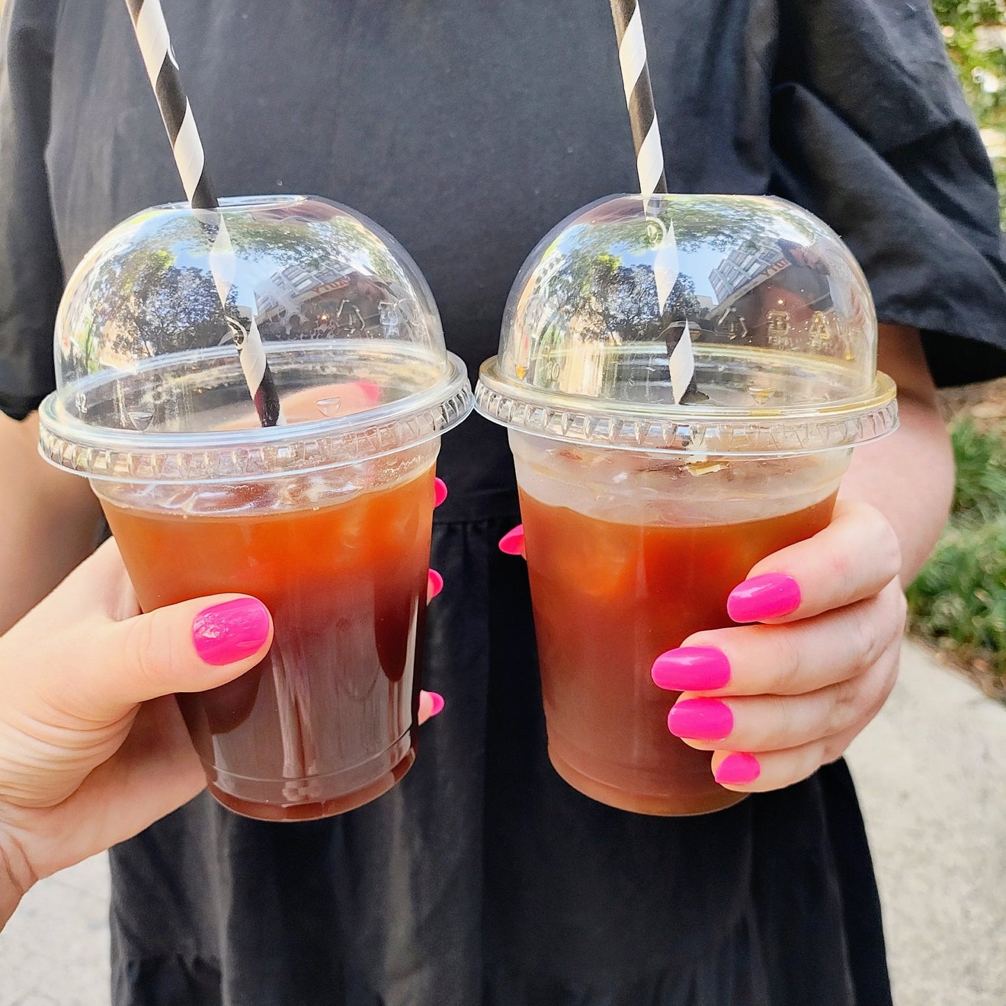 Two pale hands holding iced long blacks, you can see hot pink oval nails on the hands with a black dress in the background.