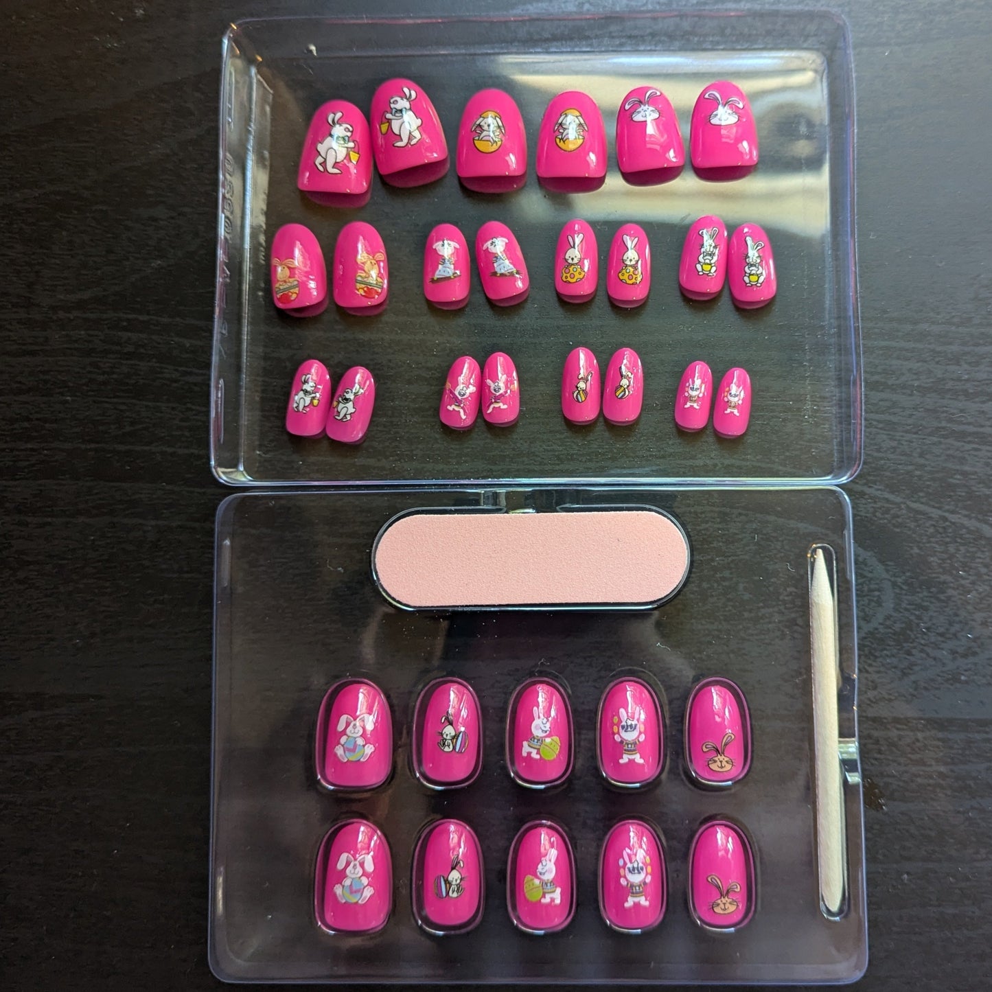 Image of a clear box with 22 oval hot pink press on gel nails with various Easter designs. Underneath is another clear box with 10 oval hot pink press on gel nails with various easter designs, a cuticle stick and nail file.