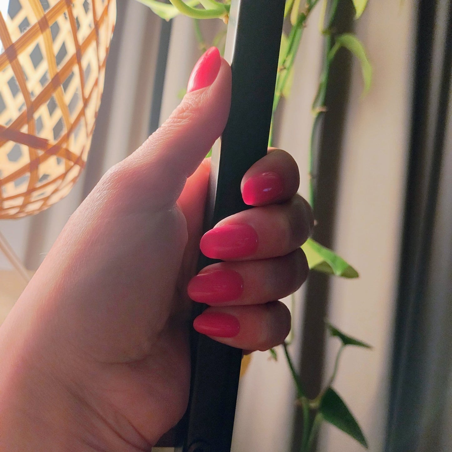 Pale left hand holding onto a black pole behind a plant and wicker light, you can see hot pink oval nails on the hand