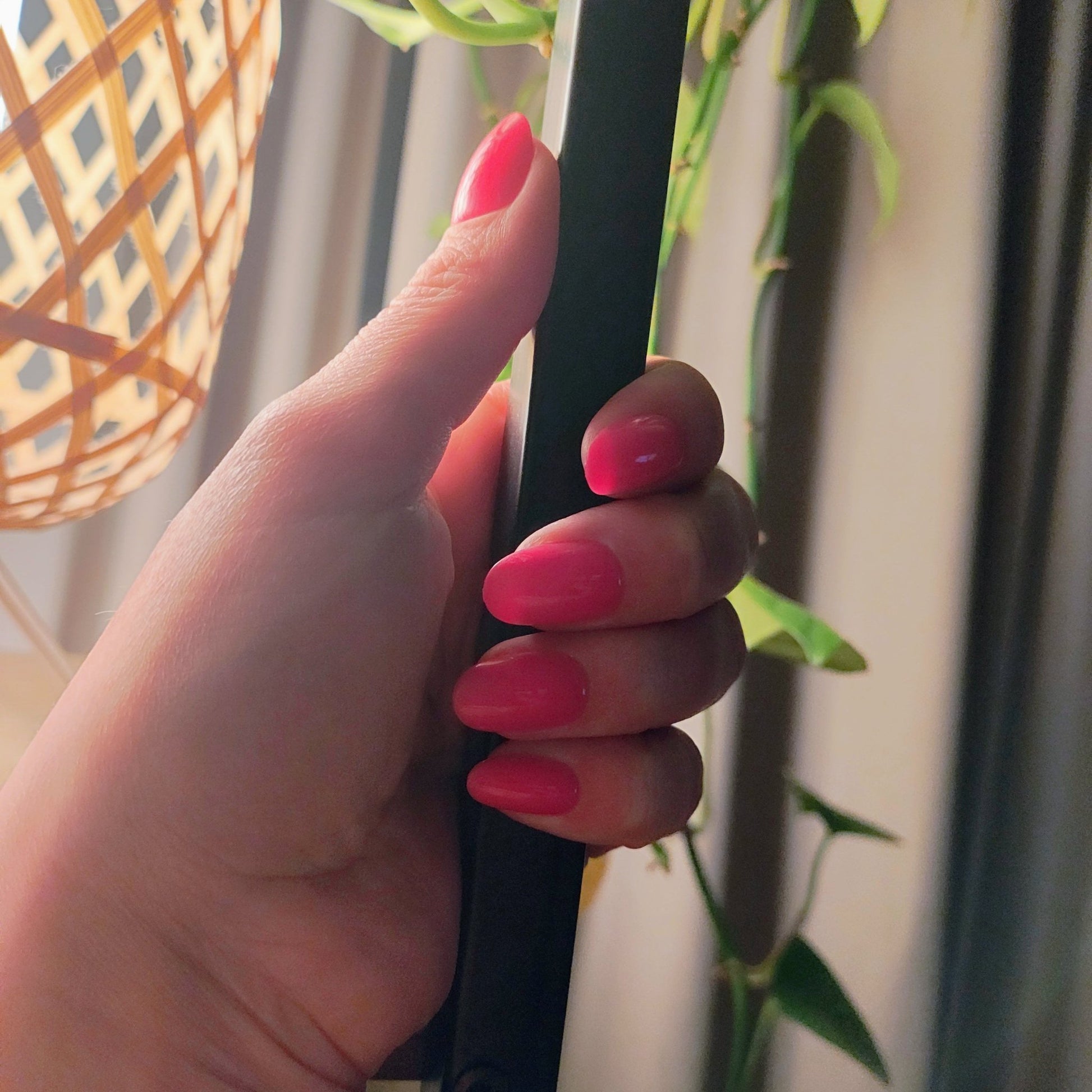 Pale left hand holding onto a black pole behind a plant and wicker light, you can see hot pink oval nails on the hand