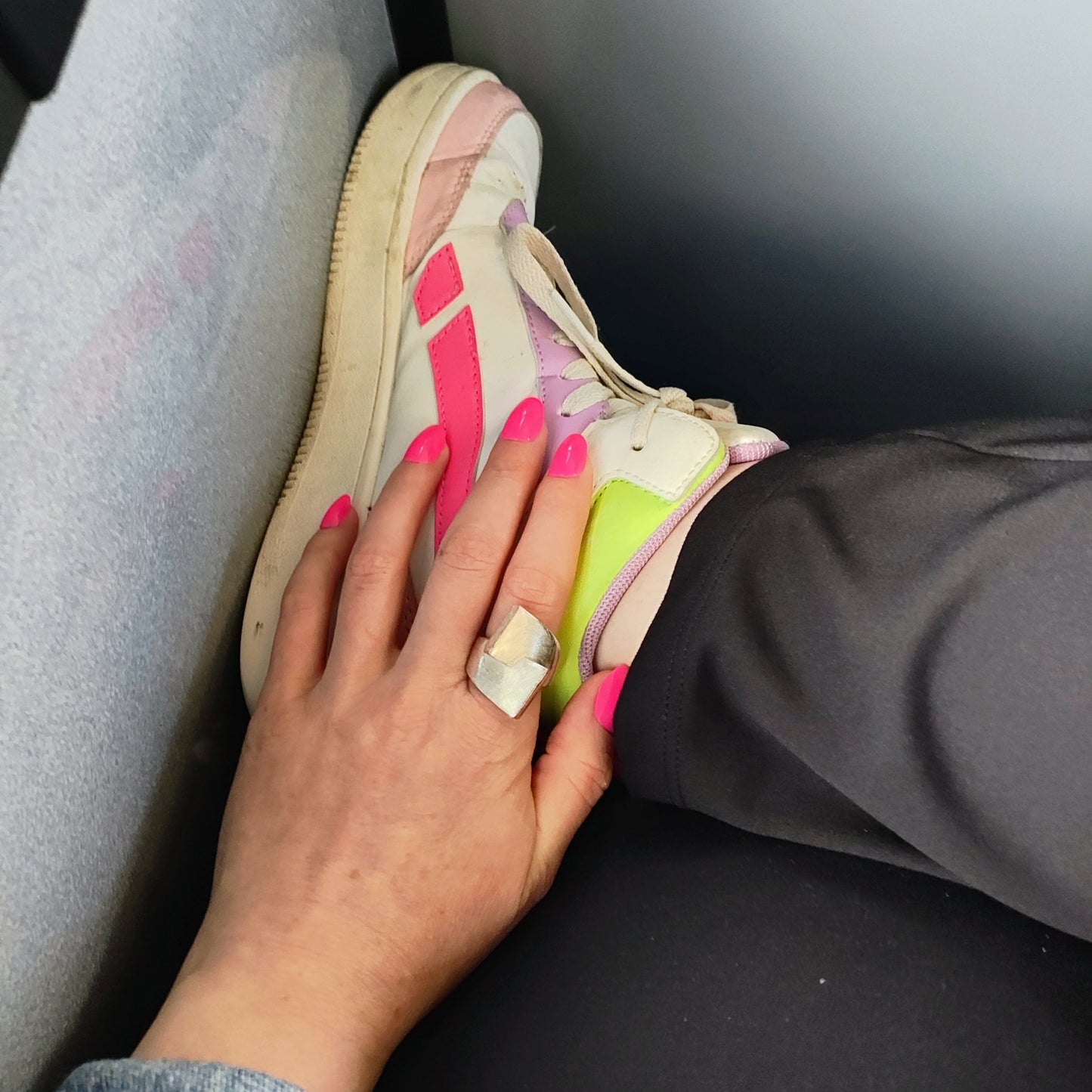 Left pale hand with hot pink oval shaped nails on top of a crossed leg with a white sneaker with pink, purple and green details.