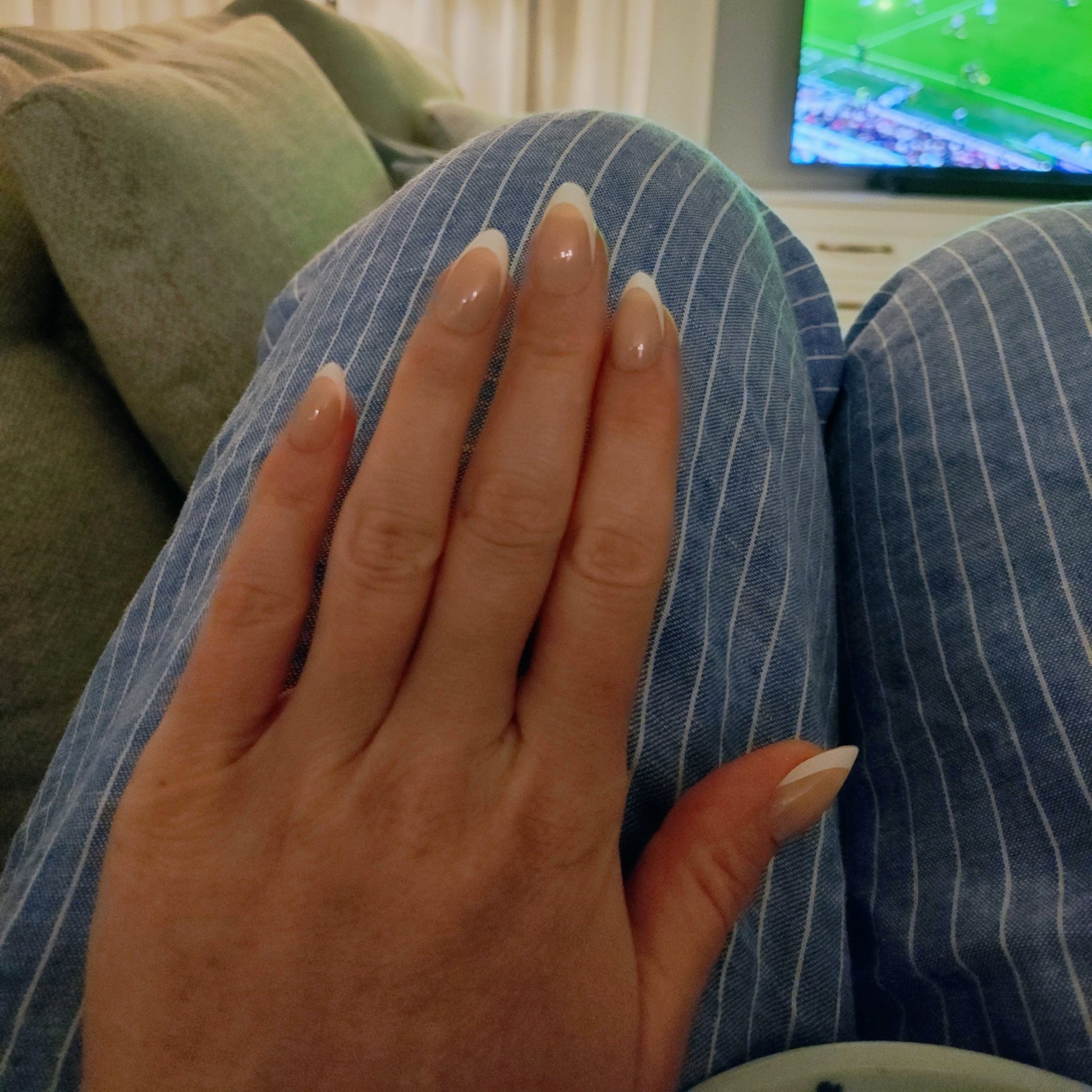 Left nail showing a classic almond French tip on a blue panted leg with a TV in the background.