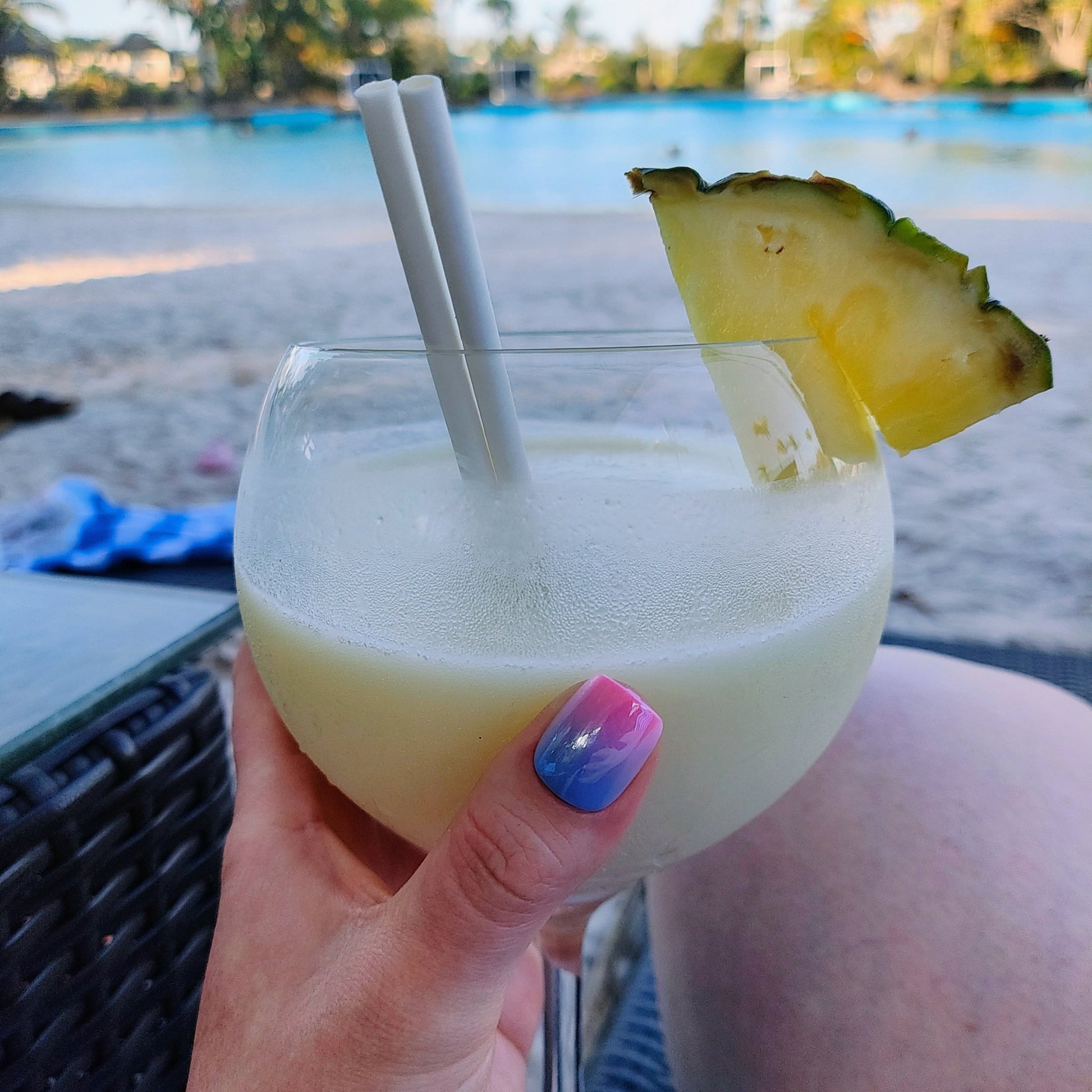 Left hand showing square nail in a metallic ombre of blue and pink holding a cocktail glass by a beach.
