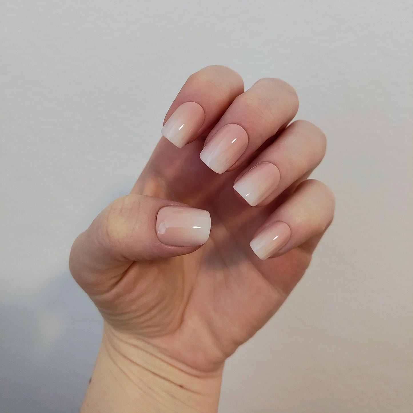 Left hand showing square nails with an ombre effect of peach to white on a white wall background.