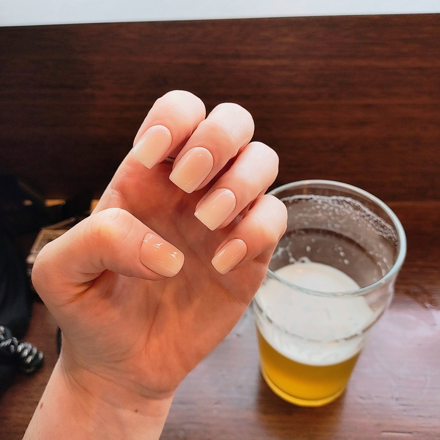 Left hand showing square nails with an ombre effect of peach to white behind a pint of beer.