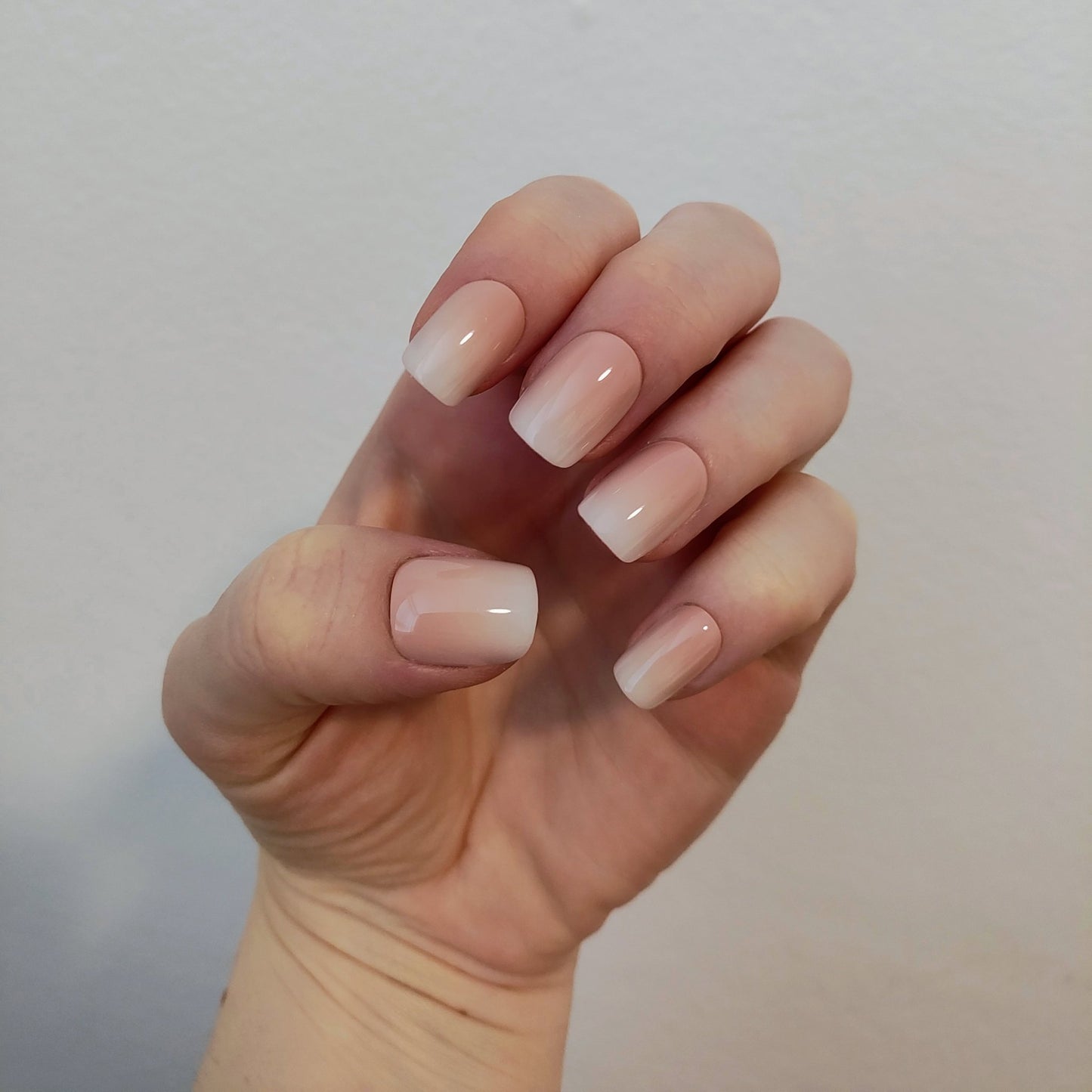 Left hand showing square nails with an ombre effect of peach to white on a white wall background.