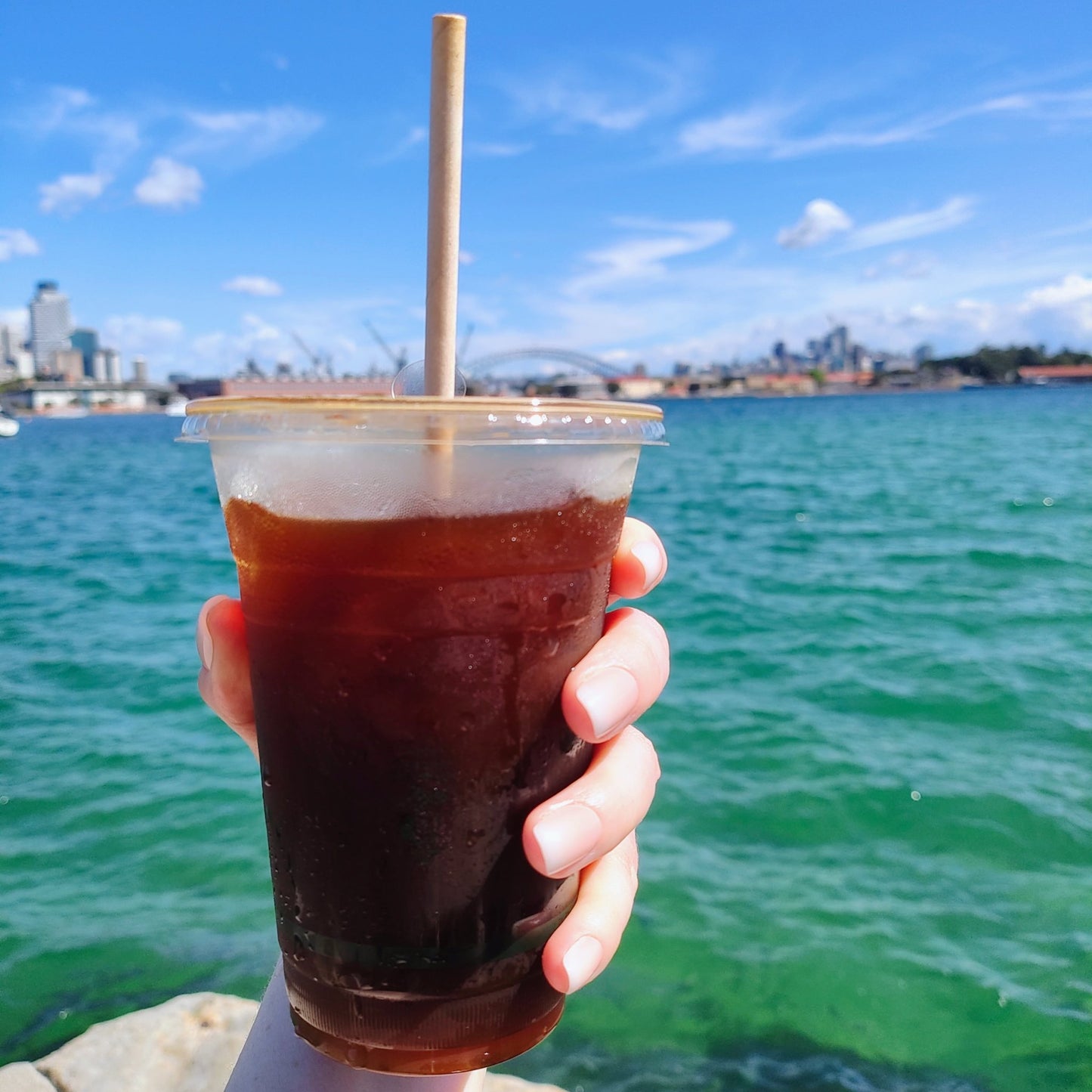 Left hand showing square nails with an ombre effect of peach to white holding an iced long back with Sydney Harbour backdrop.
