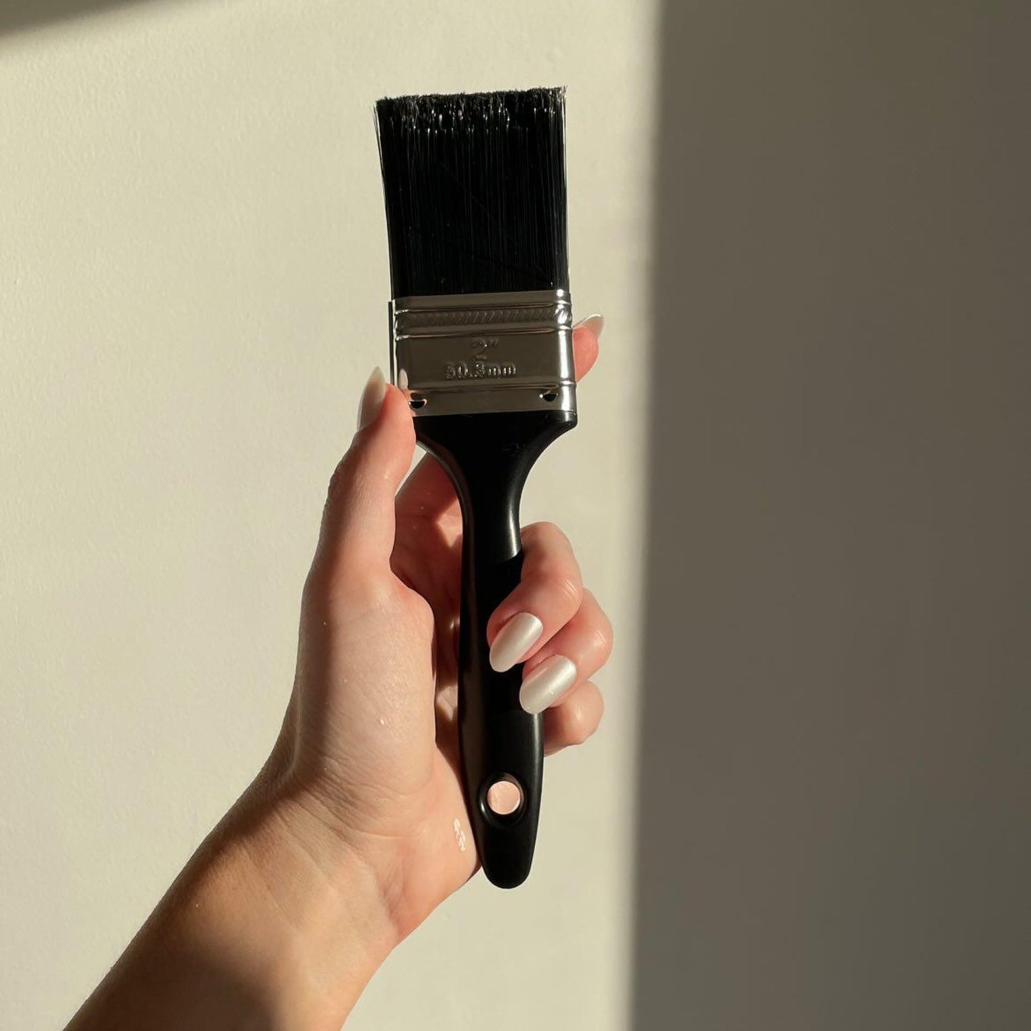 Left hand showing oval pearl coloured nails holding a paint brush on a white wall.