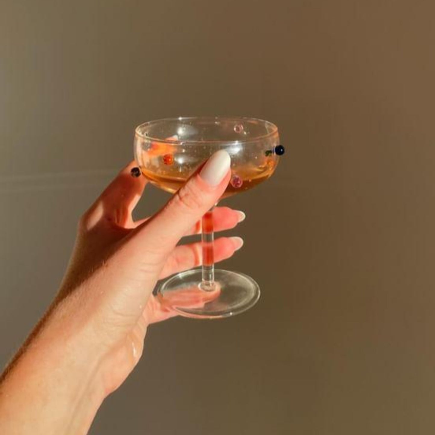 Left hand showing oval pearl coloured nails holding a cocktail glass on a cream wall.