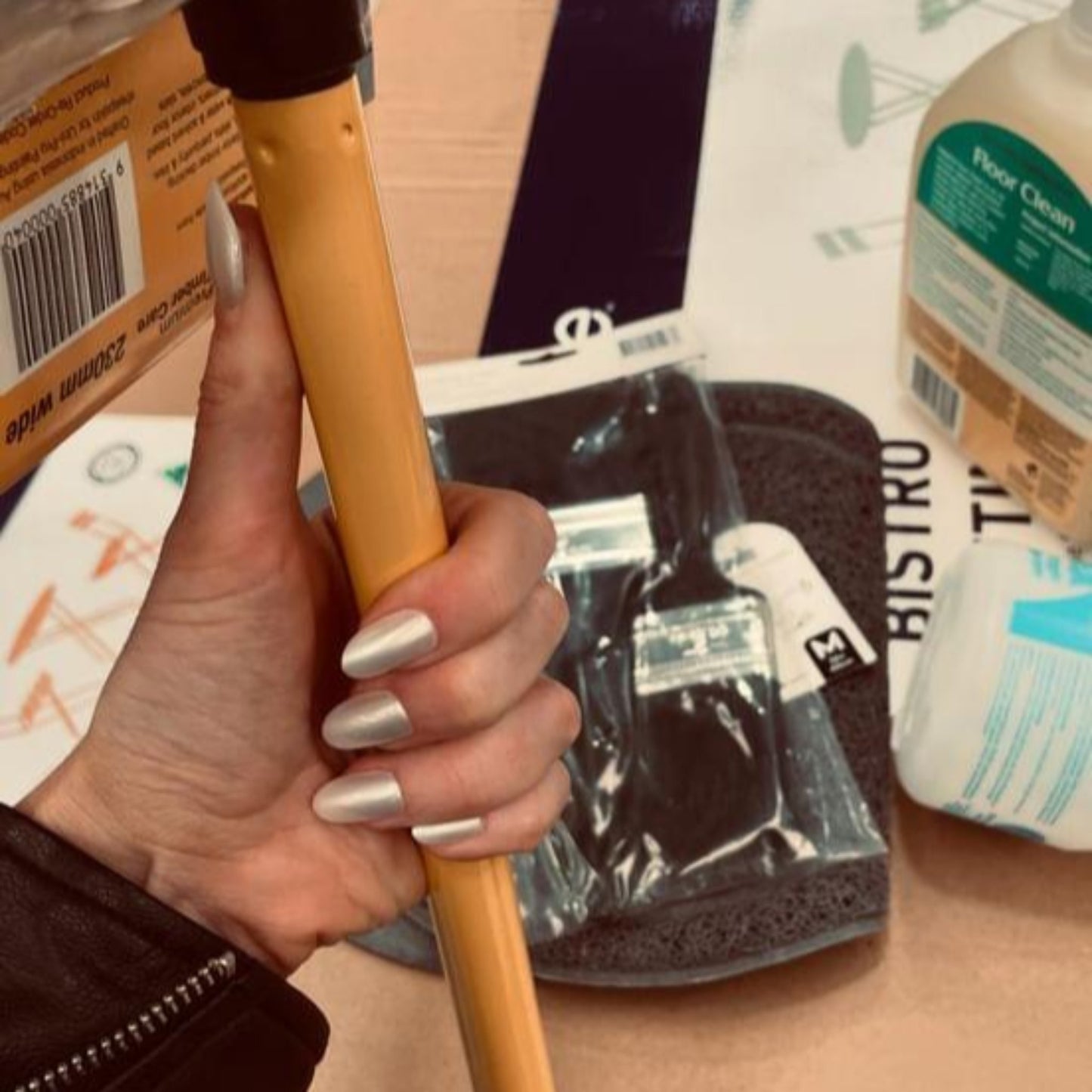 Left hand wearing oval pearl coloured nails holding a broom in a hardware store.