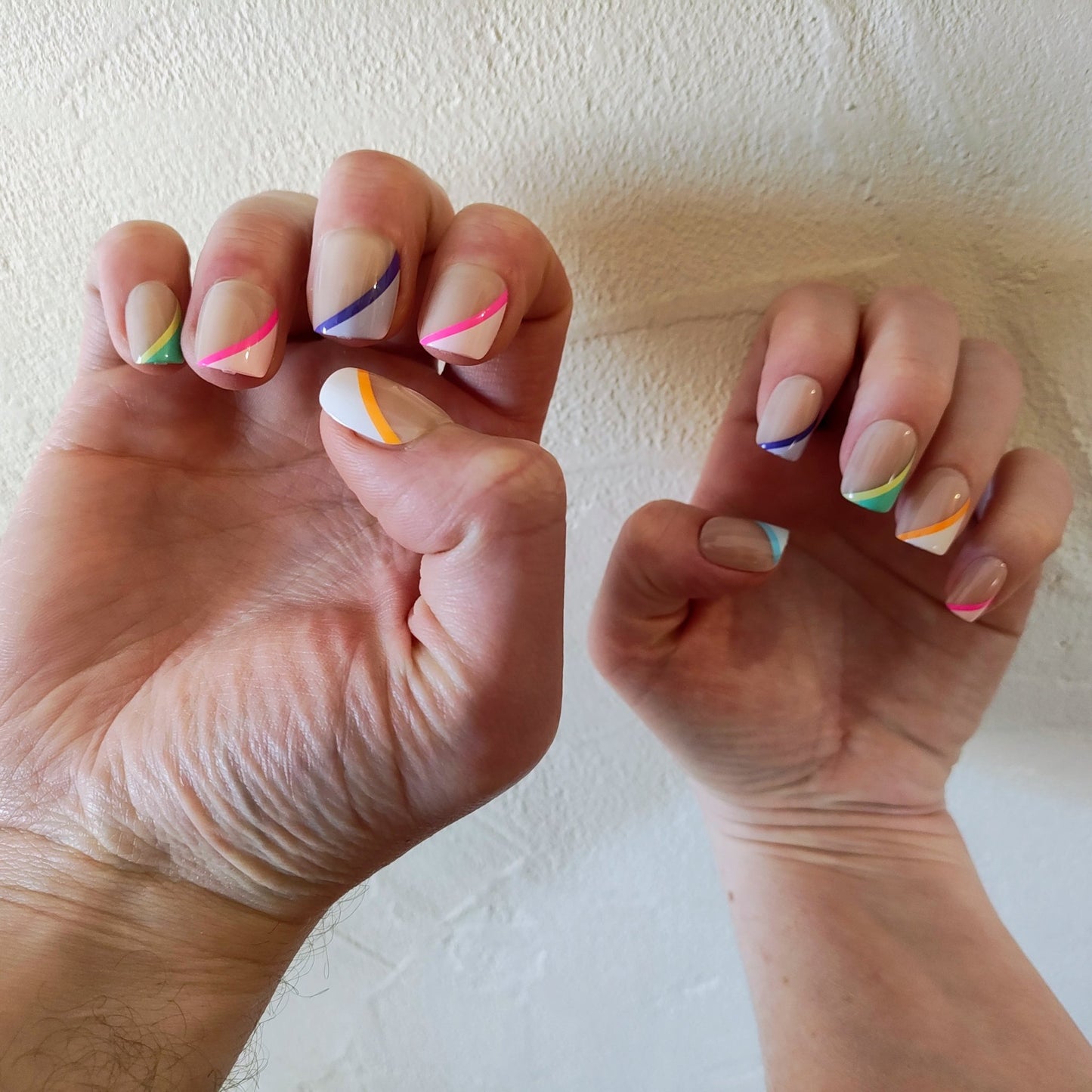 Two hands with square cut nails with angular colour stripes, the left one larger than the right behind a cream wall. The colours of the stripes are: Left hand - green, pink, purple, pink and orange. Right hand - blue, purple, green, orange and pink.