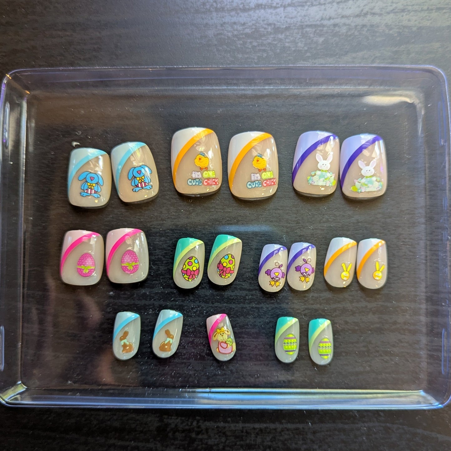 A clear box with square gel press-ons with 19 nails of varying tip colours and Easter designs.