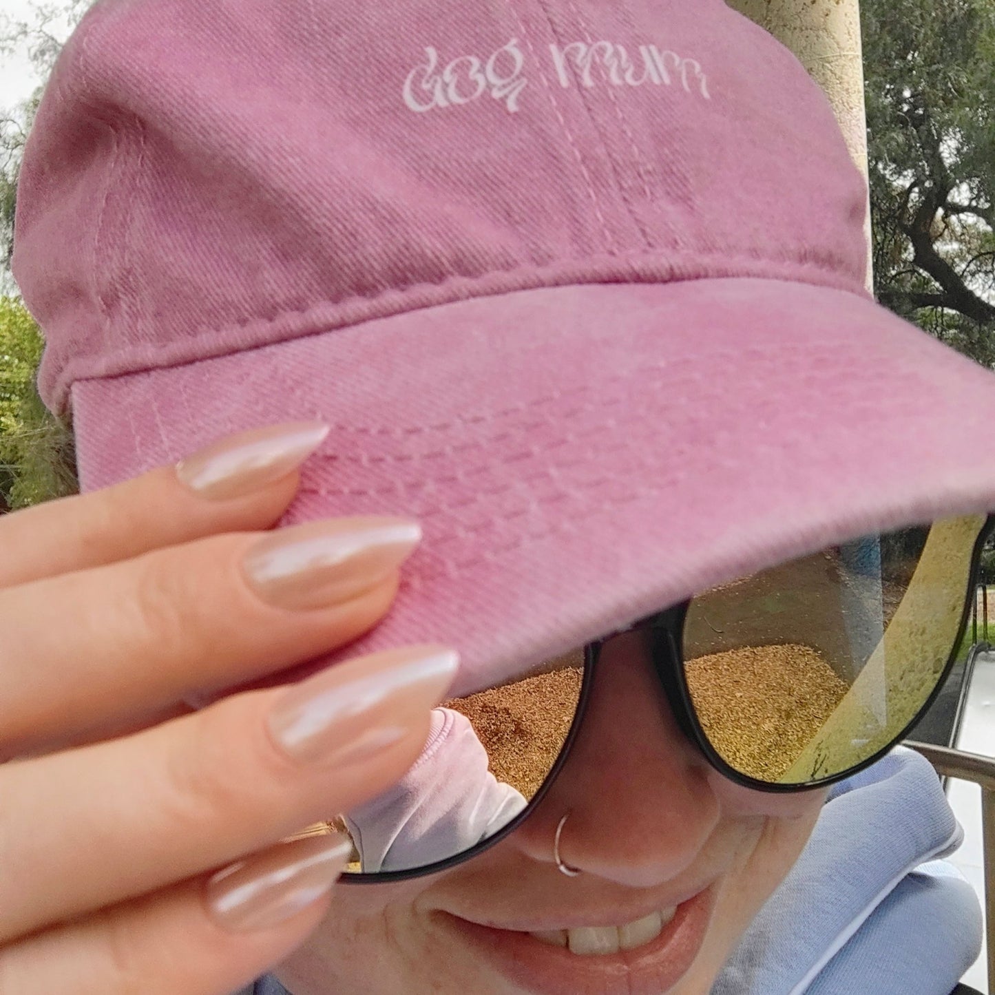 Left hand showing an almond shimmery, pinky peach coloured nails on a woman wearing a pink 'dog mum' hat.