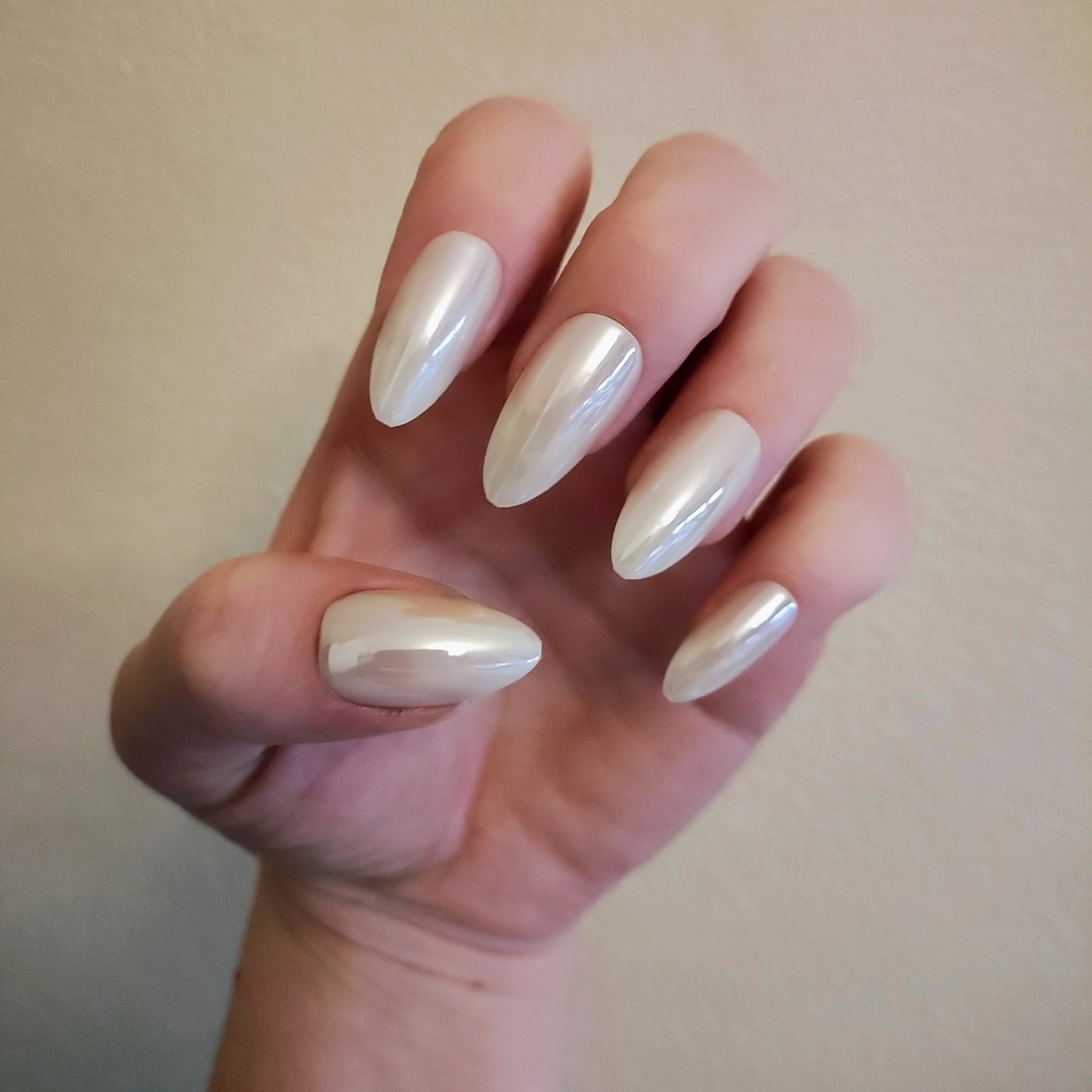 Left hand with long almond white pearl frosted nails in front of cream wall.