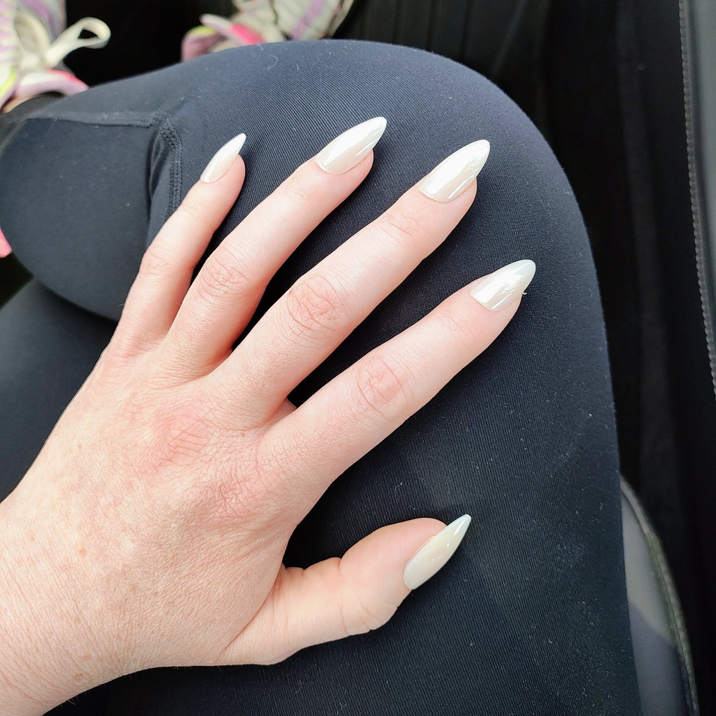 Left hand with long almond white pearl frosted nails on top of crossed legs wearing black leggings.