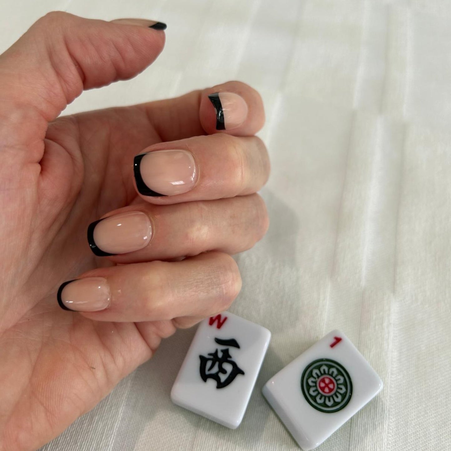 Left hand showing square nails with a black French tip on next to a Mahjong tiles.