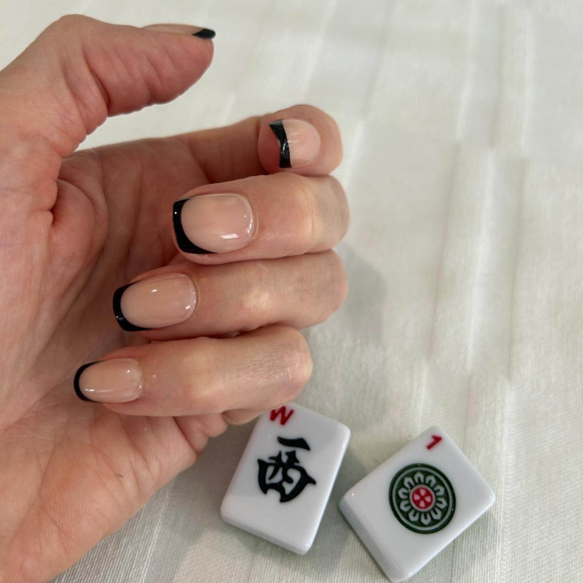 Left hand showing square nails with a black French tip on next to a Mahjong tiles.