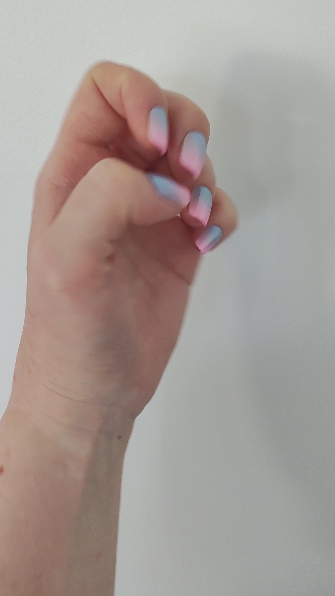 Left hand showing square nails in a metallic ombre of blue and pink against a white wall.