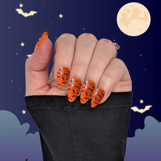 A left hand wearing medium long almond nails in pumpkin orange with small black bat details on a halloween background.