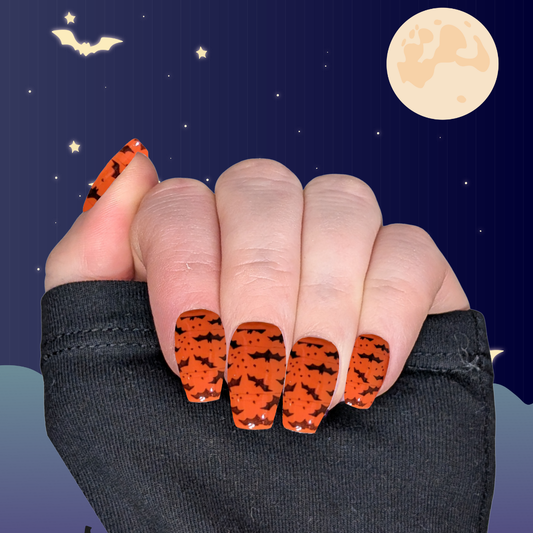 A left hand wearing medium long coffin nails in pumpkin orange with small black bat details on a halloween background.