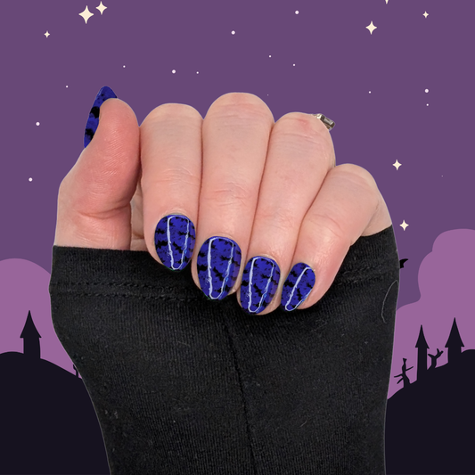 A left hand wearing XS almond nails in a deep purple with small black bat details on a halloween background.