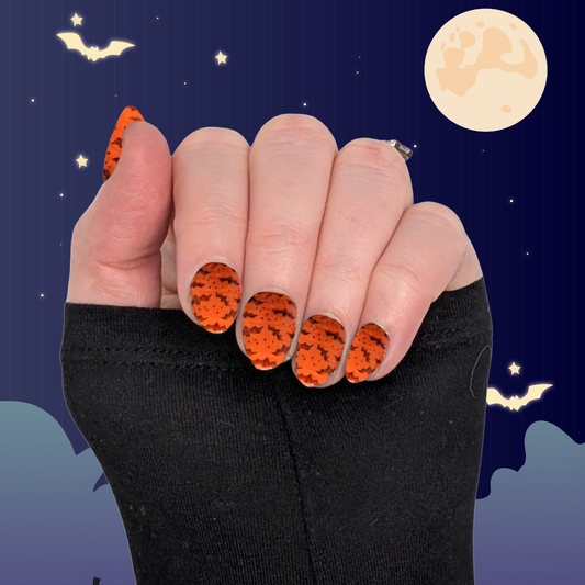 A left hand wearing XS almond nails in pumpkin orange with small black bat details on a halloween background.