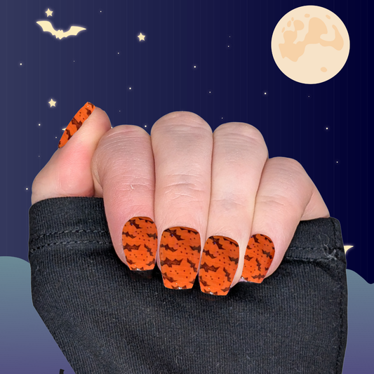 A left hand wearing XS coffin nails in pumpkin orange with small black bat details on a halloween background.