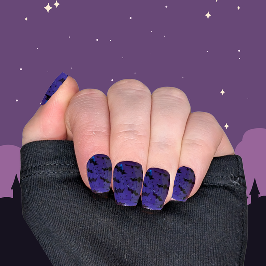 A left hand wearing XS coffin nails in witchy purple with small black bat details on a halloween background.