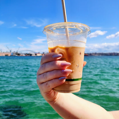 Right hand holding an iced latte showing latte brown in our almond shape, with a slight shimmer, Sydney harbour in the background.