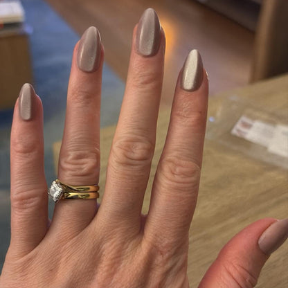 Left hand wearing almond shaped nails in a shiny chocolatey colour, with a lounge room in the background. 