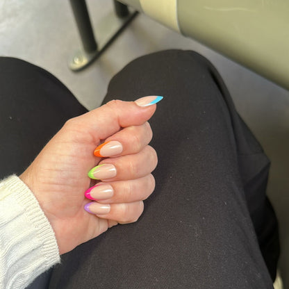 Left hand on a pair of black pants on a train. Almond french tip multi coloured tips: blue, orange, green, pink and purple.