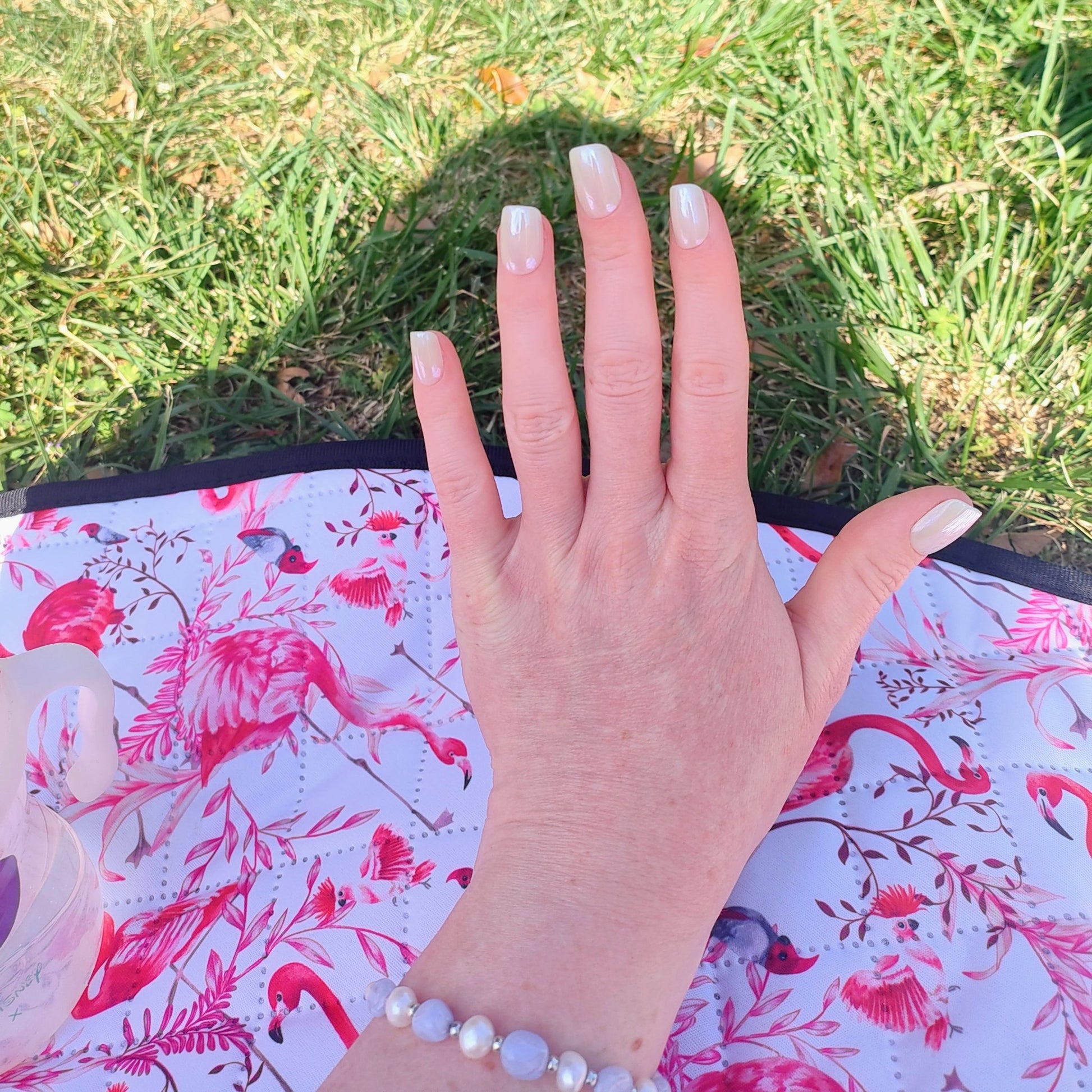 Left hand showing square pearl coloured nails on a pink and white picnic rug.