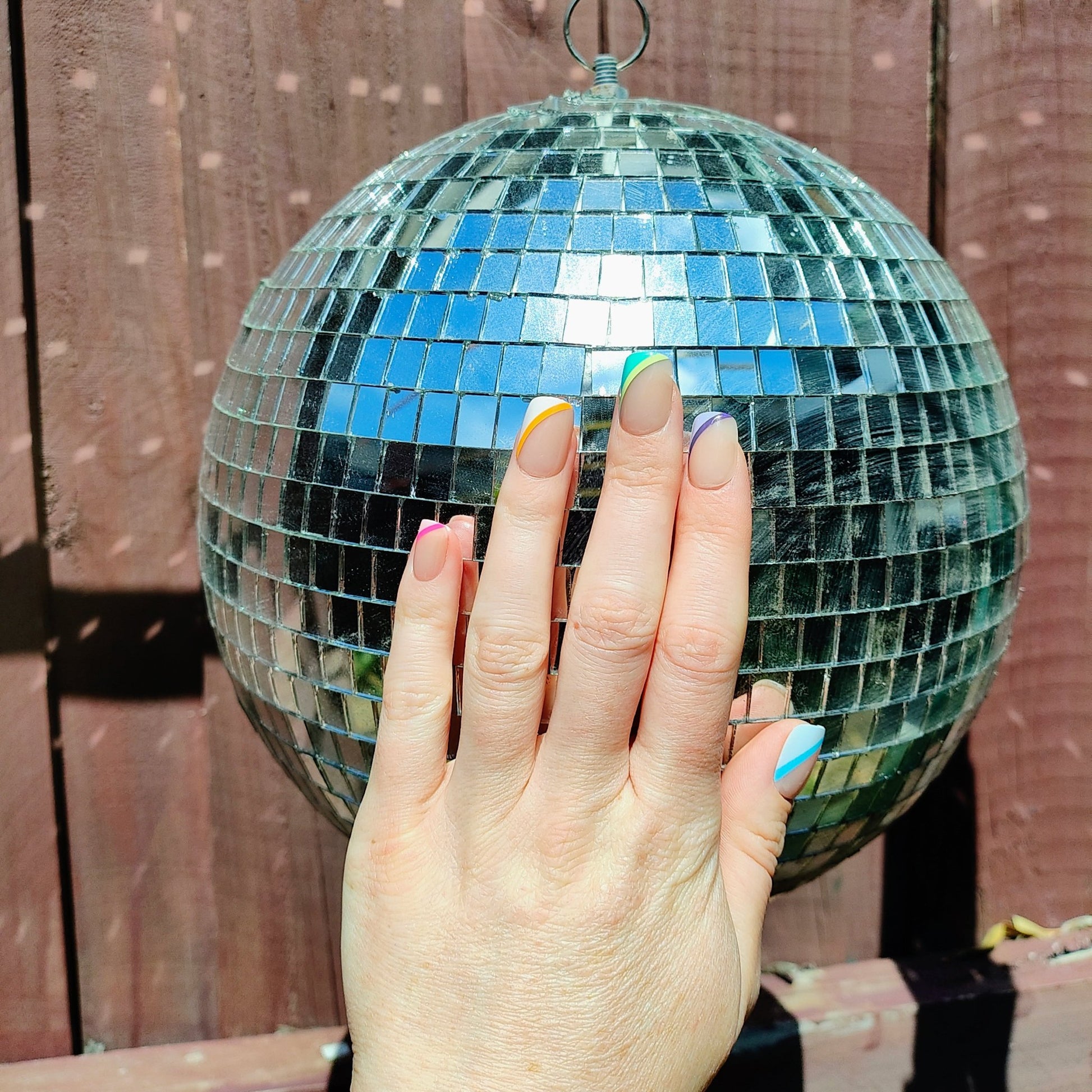 Square cut nails on left hand with striped angled stripes on left hand side of each nail behind a disco ball. The stripes are pink, orange, green, purple and blue.