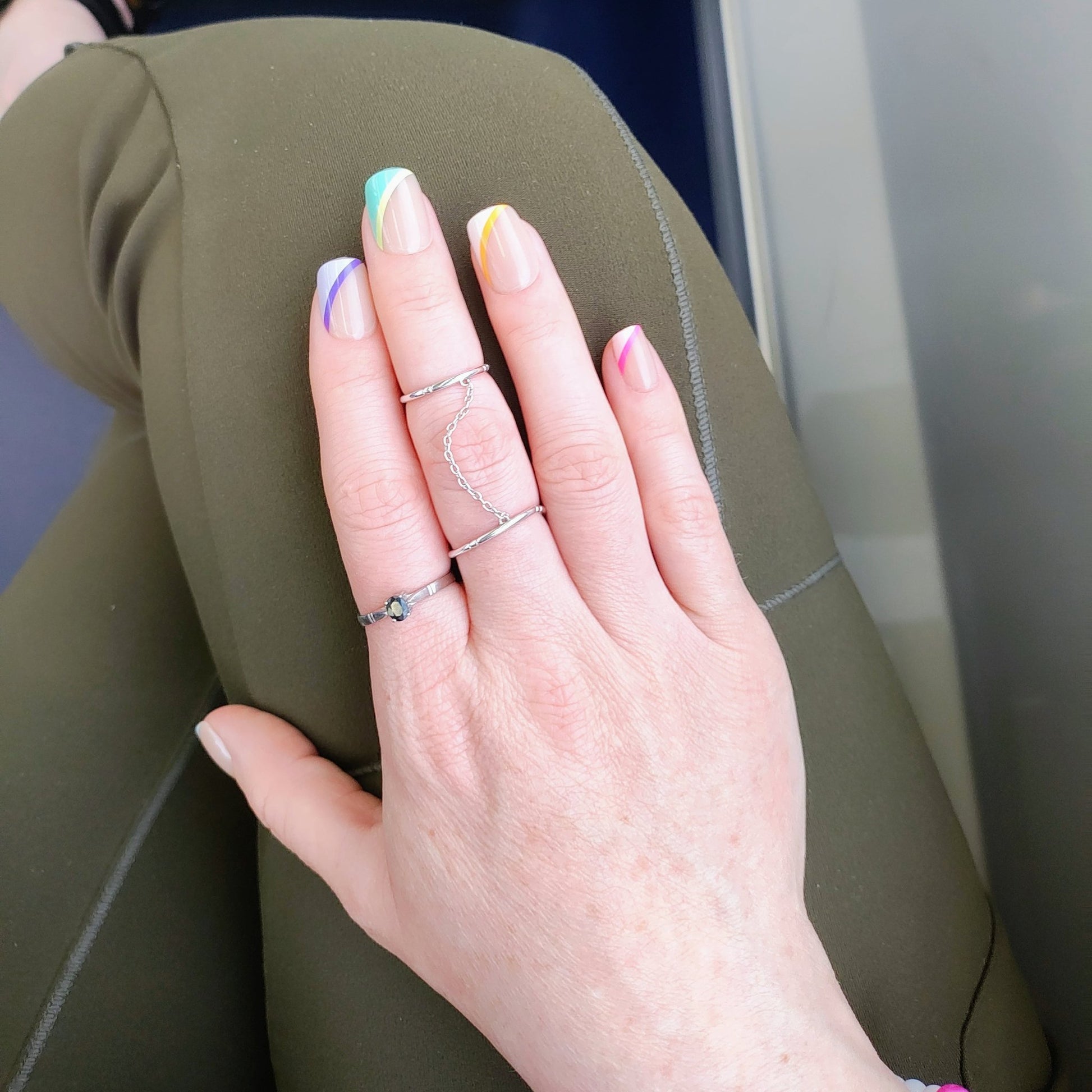 Square cut nails on right hand with striped angled stripes on left hand side of each nail on a leg sitting on a Melbourne train. The stripes are blue, purple, green, orange and pink. 