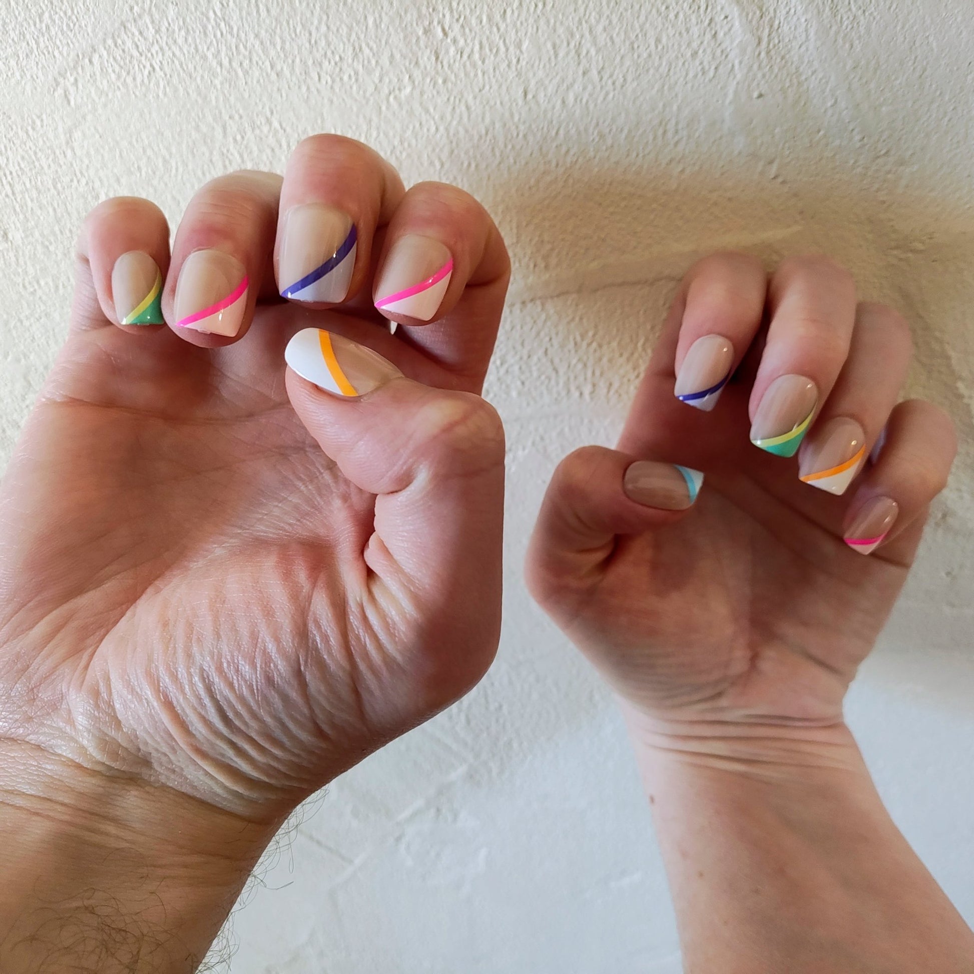 Two hands with square cut nails with angular colour stripes, the left one larger than the right behind a cream wall. The colours of the stripes are: Left hand - green, pink, purple, pink and orange. Right hand - blue, purple, green, orange and pink. 