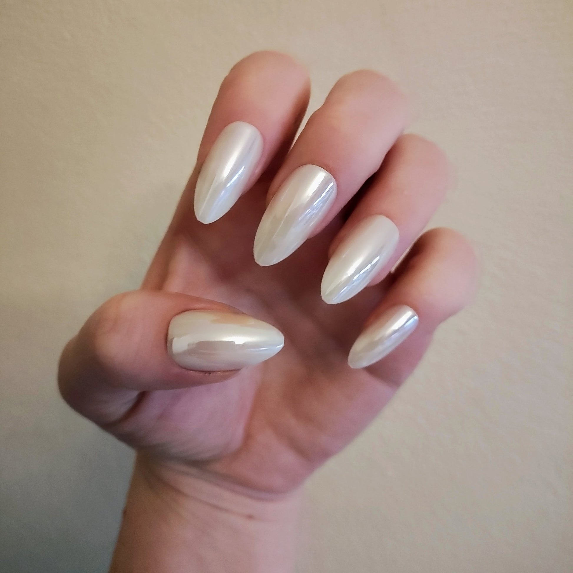 Left hand with long almond white pearl frosted nails in front of cream wall.