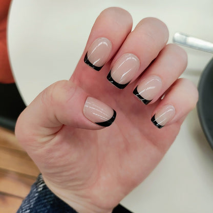 Left hand showing square nails with a black French tip on a cafe table.