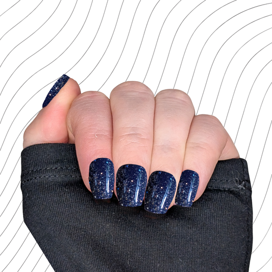 Left hand showing navy blue extra short coffin nails with a glitter gloss finish on a wavy background.