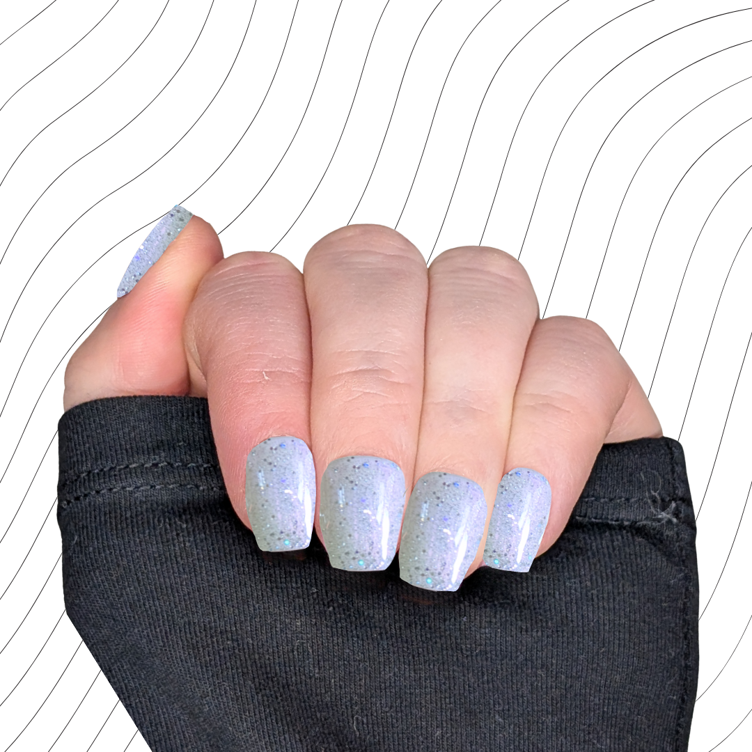 Left hand showing pearlescent white extra short coffin nails with a glitter gloss finish on a wavy background.