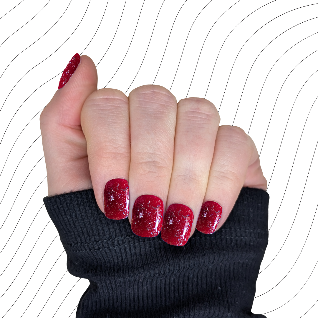 Left hand showing deep red extra short square nails with a glitter gloss finish on a wavy background.