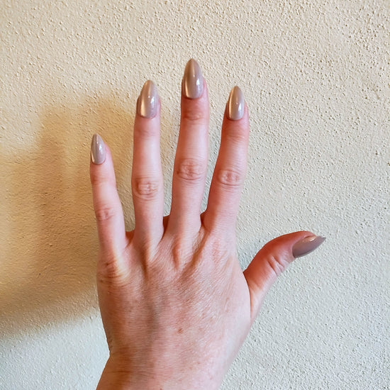 Left hand showing latte brown in our almond shape, with a slight shimmer, on a cream wall background.